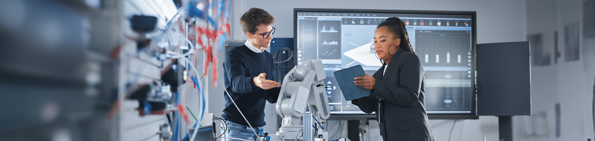 Zwei Fachkräfte arbeiten in einem technischen Labor an einem Roboterarm, während sie Daten auf einem Tablet und einem großen Bildschirm auswerten.