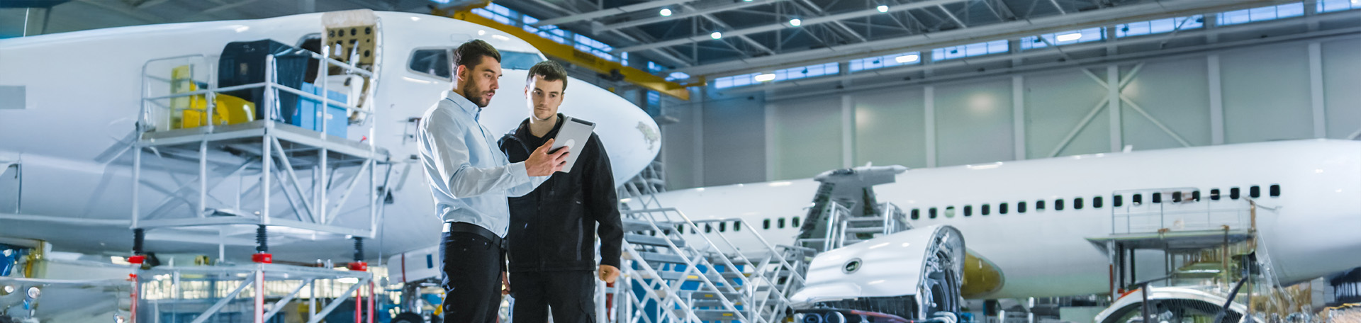 Zwei Fachkräfte überprüfen in einer Flugzeughalle ein Flugzeug und besprechen technische Informationen anhand eines Tablets.